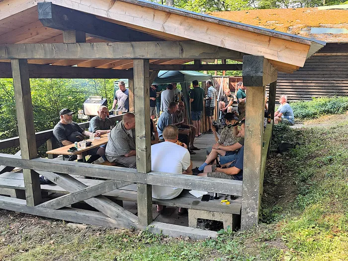 Eine Gruppe von etwa 20 Männern sitzt und steht unter einem Holzpavillon, einige essen und trinken an Tischen. Im Hintergrund sind weitere Personen und Büsche zu sehen.