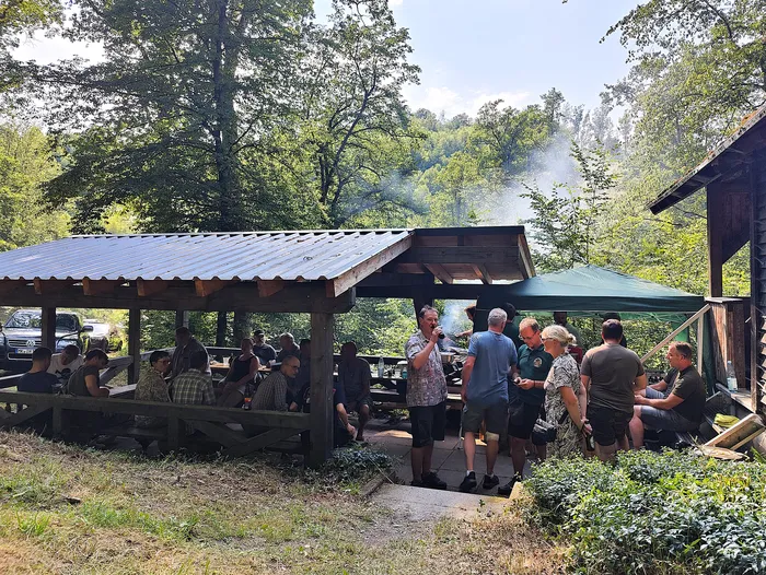 Menschen sitzen und stehen unter einem überdachten Holzbalken zusammen, einige essen und unterhalten sich. Im Hintergrund sind Bäume und ein Auto zu sehen.