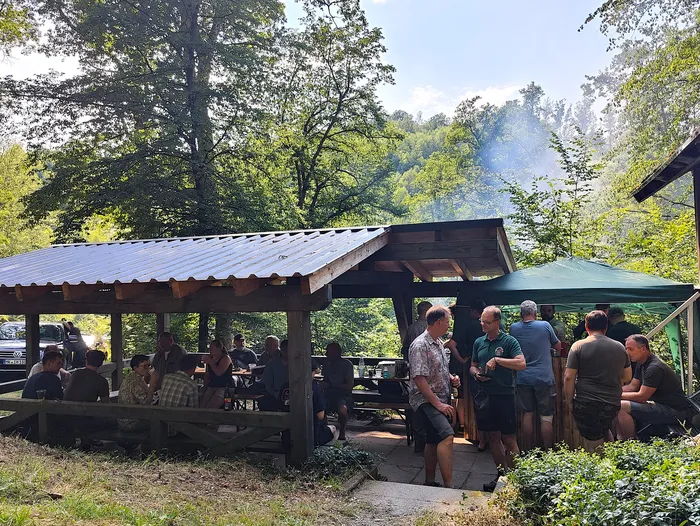Menschen sitzen und stehen unter einem überdachten Holzbogen zusammen, einige essen und unterhalten sich. Im Hintergrund sind Bäume und ein Auto zu sehen.