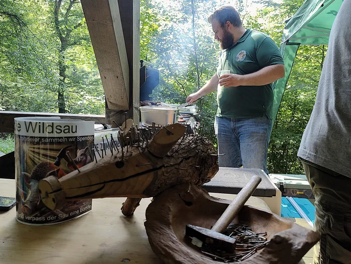 Ein Mann steht an einem Grill und hält ein Bratenschaufel bereit. Im Vordergrund steht eine Holzskulptur eines Wildschweins und ein Werbebecher für Spendenaktionen des Hegering Schalksmühle-Hülscheid.