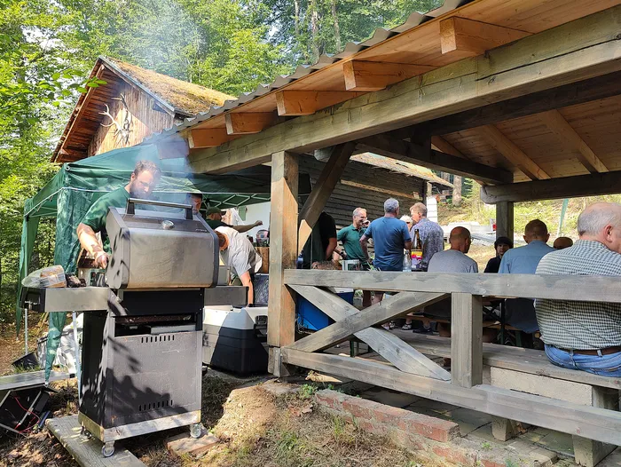 Ein Mann steht an einem großen Grill unter einem Holzpavillon und bereitet Speisen zu, während mehrere Personen an Tischen sitzen und essen. Im Hintergrund ist ein Wald zu sehen.