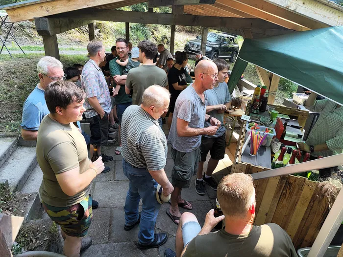 Eine Gruppe von Männern steht unter einem Holzpavillon und unterhält sich. Ein Mann bedient an einem improvisierten Ausschank mit Getränken und Essensutensilien. Im Hintergrund ist ein schwarzer Geländewagen geparkt.