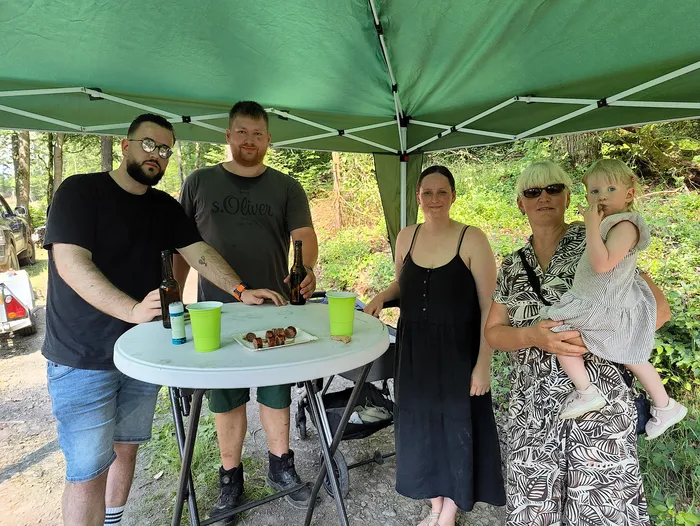 Fünf Personen stehen um einen Tisch unter einem grünen Pavillon. Auf dem Tisch stehen Getränke und ein Teller mit kleinen Fleischstücken. Im Hintergrund ist ein Wald zu sehen.