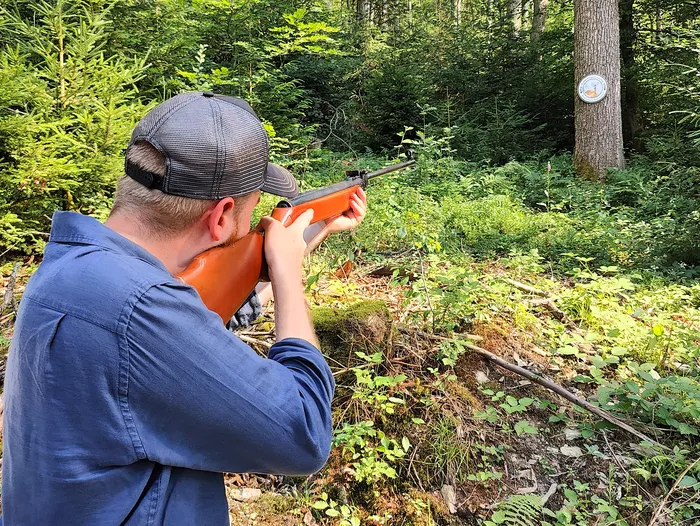 Ein Mann zielt mit einem Gewehr in einem Wald. Im Hintergrund ist ein Zielscheibe an einem Baumstamm zu sehen.