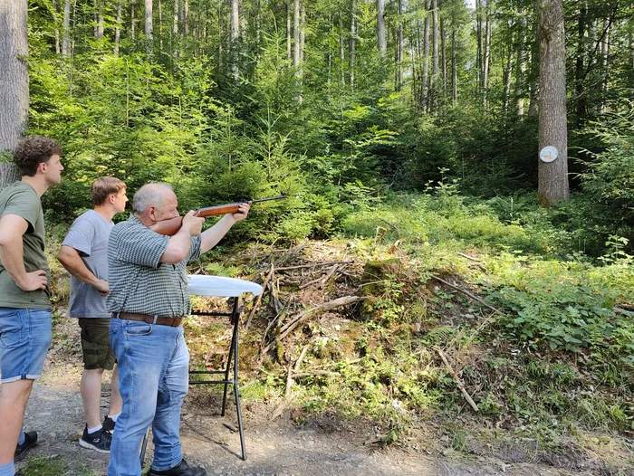 Ein Mann zielt mit einem Gewehr auf eine Zielscheibe in einem Wald, während zwei weitere Männer ihm zusehen. Im Hintergrund sind Bäume und dichtes Unterholz zu erkennen.