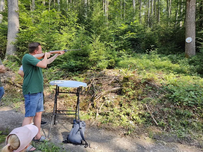 Ein Mann zielt mit einem Gewehr auf eine Zielscheibe, die auf einem Tisch steht. Im Hintergrund ist dichter Wald zu sehen, und weitere Personen sind teilweise erkennbar.