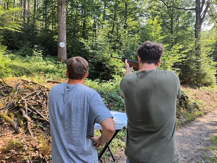 Ein Mann zielt mit einem Gewehr auf eine Zielscheibe im Wald, während ein zweiter Mann daneben steht und zusieht. Im Hintergrund ist dichter Wald zu erkennen.