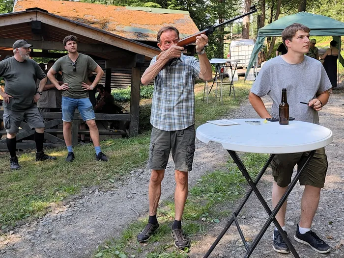 Ein Mann zielt mit einem Gewehr auf eine Zielscheibe, während andere Teilnehmer des Jägerpicknicks zusehen. Im Hintergrund steht ein Tisch mit Getränken und ein Zelt.