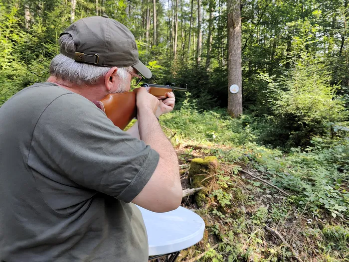 Ein Mann zielt mit einem Gewehr im Wald. Im Hintergrund ist eine Zielscheibe an einem Baumstamm zu erkennen.