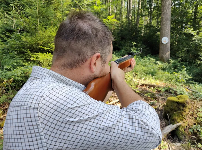 Ein Mann zielt mit einem Gewehr im Wald. Im Hintergrund ist eine Zielscheibe an einem Baumstamm zu erkennen.