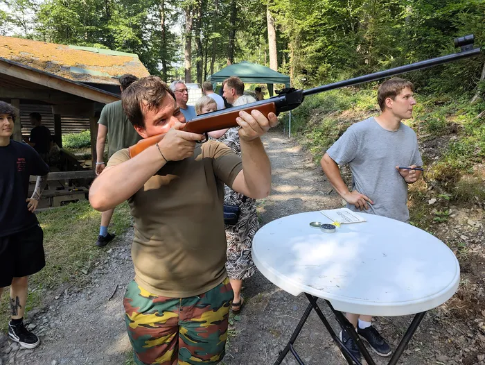 Ein Mann zielt mit einem Gewehr auf eine Zielscheibe, während andere Teilnehmer des Jägerpicknicks zusehen. Im Hintergrund sind Bäume und ein Gebäude zu erkennen.