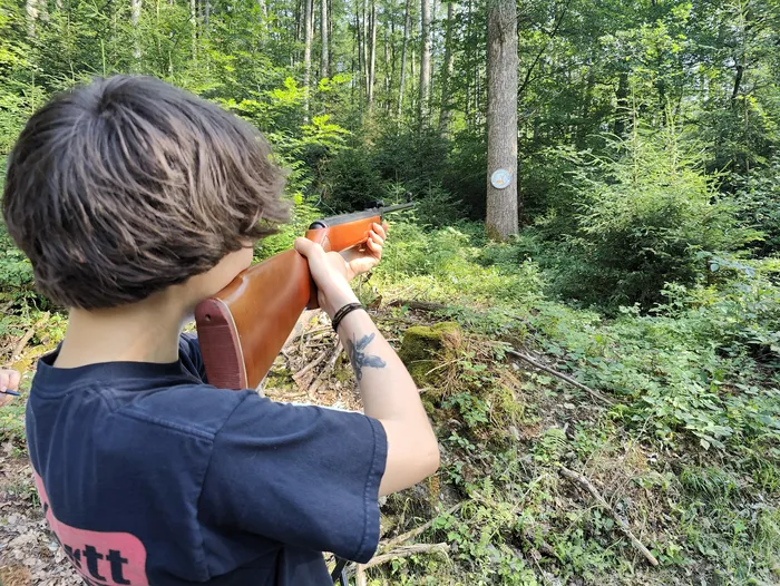 Eine Person zielt mit einem Gewehr in einem Wald. Im Hintergrund sind Bäume und Büsche zu sehen, rechts ein Zielscheibe.