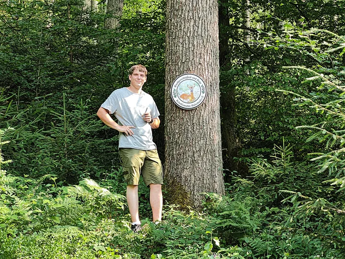 Junger Mann steht lächelnd neben einem dicken Baumstamm mit einem Schild, das ein Reh zeigt. Im Hintergrund dichter Wald.