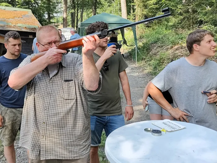 Ein Mann zielt mit einem Gewehr, während ein anderer ihn dabei unterstützt. Im Hintergrund stehen weitere Personen und ein Tisch mit Zubehör.