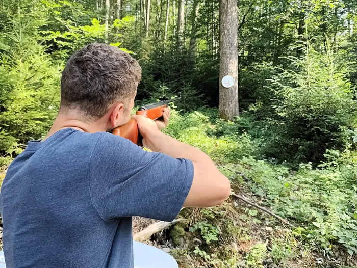 Ein Mann zielt mit einem Gewehr in einem Wald. Im Hintergrund ist eine Zielscheibe an einem Baumstamm zu sehen.
