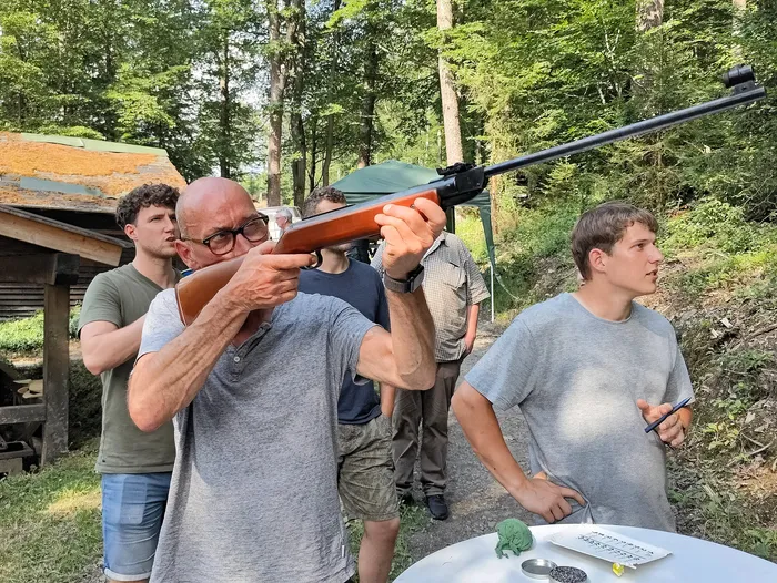 Ein Mann zielt mit einem Gewehr, während zwei weitere Personen ihn dabei unterstützen und beobachten. Im Hintergrund steht ein Gebäude und Bäume. Ein Tisch mit Zielscheiben und einer kleinen Figur ist im Vordergrund zu sehen.