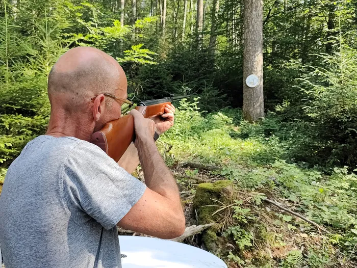 Ein Mann zielt mit einem Gewehr in einem Wald. Im Hintergrund sind Bäume und dichtes Grün zu sehen.