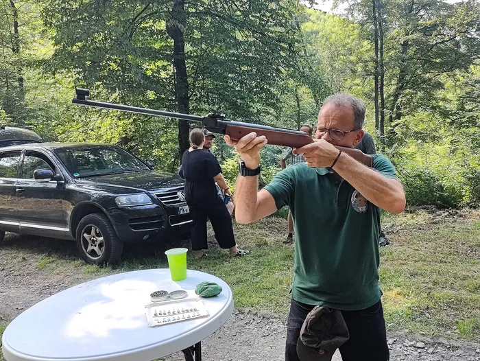 Ein Mann zielt mit einem Gewehr im Wald. Im Hintergrund steht ein schwarzer Geländewagen und eine weitere Person. Auf einem Tisch im Vordergrund befinden sich Munition und ein Getränk.