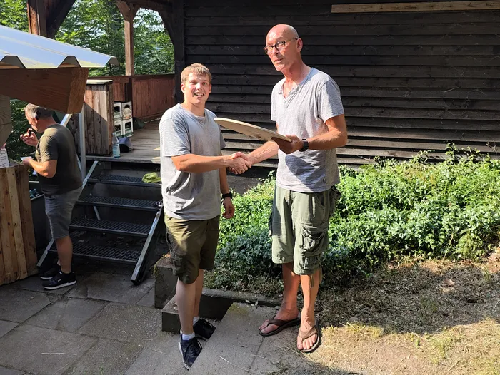 Zwei Männer geben sich die Hand, einer von ihnen hält eine Holztafel. Im Hintergrund steht ein Mann an einem Holztisch und ein Gebäude mit einem Satteldach.
