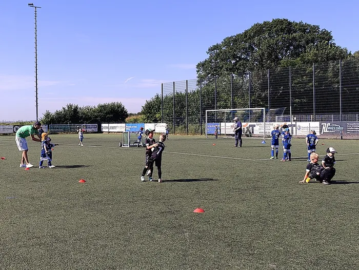 Fußballtraining mit Kindern auf einem grünen Kunstrasenplatz. Mehrere Spieler in Trikots stehen und laufen, während ein Trainer Anweisungen gibt. Im Hintergrund ein Fußballtor und Werbebanner.