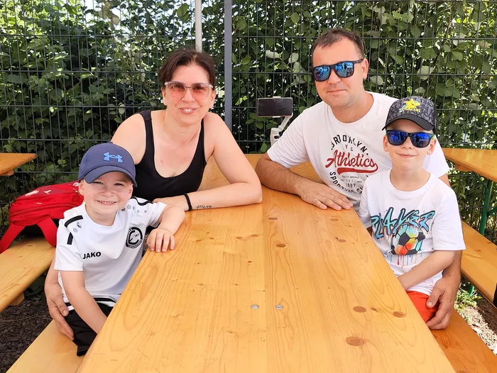 Eine Familie mit zwei Kindern sitzt an einem Holztisch im Freien. Die Eltern tragen Sonnenbrillen, die Kinder Baseballkappen. Im Hintergrund ist ein grüner Zaun zu sehen.