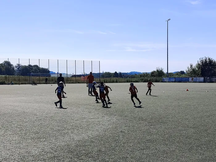 Eine Gruppe Jugendlicher in Fußballtrikots spielt auf einem Hartplatz. Im Hintergrund ein Zaun, Bäume und ein Mast mit Flutlicht.