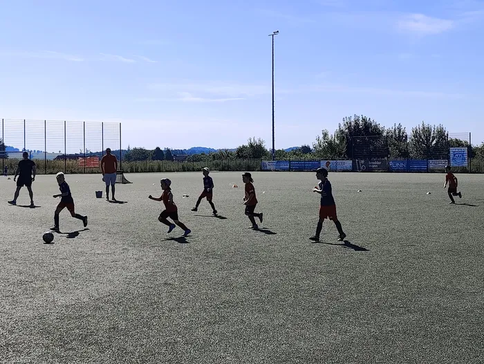 Kinder spielen Fußball auf einem Kunstrasenplatz. Im Hintergrund steht ein Zaun und Bäume, sowie Werbebanner.