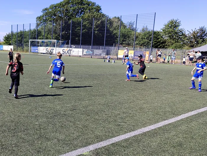 Junge Fußballspieler in blauen und roten Trikots laufen auf einem grünen Kunstrasenplatz. Im Hintergrund sind Zuschauer und Werbebanner zu sehen.