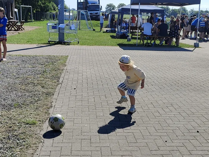 Ein Kind mit blonden Locken und einer weißen Kappe schießt auf einem gepflasterten Platz einen Fußball. Im Hintergrund sind weitere Menschen und Zelte zu sehen.