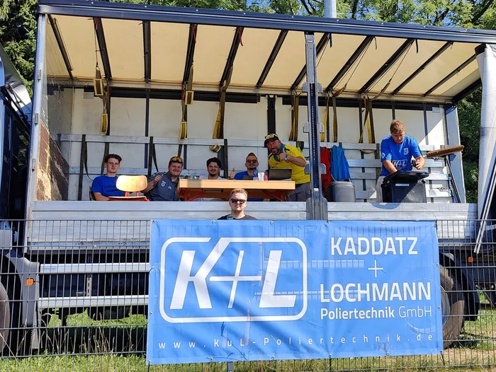 Sechs Personen sitzen oder stehen auf einer erhöhten Plattform unter einem weißen Dach. Im Vordergrund ein blaues Banner mit dem Aufdruck 