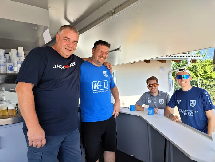 Vier Männer stehen lächelnd vor einem mobilen Getränkestand mit Bierfässern. Zwei von ihnen tragen T-Shirts mit dem Logo des Sponsors K+L, die anderen Sportbekleidung mit Vereinslogo.