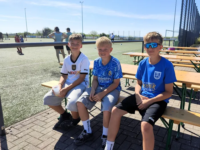 Drei Jungen sitzen auf einer Bank vor einem Fußballfeld und essen oder trinken. Im Hintergrund sind weitere Spieler und Zuschauer zu sehen.