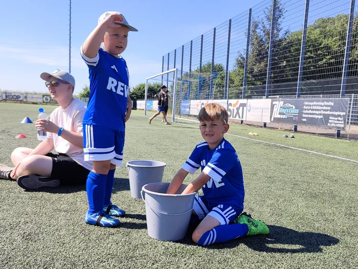 Zwei junge Fußballspieler in blauen Trikots und Stutzen knien auf einem Kunstrasenplatz und halten Eimer bereit. Im Hintergrund steht ein Trainer und weitere Spieler trainieren.