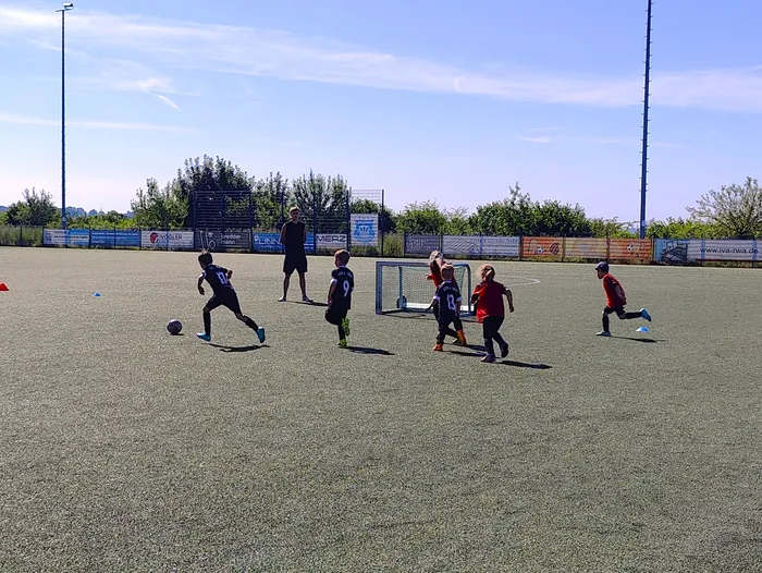 Fußballspieler in bunten Trikots während eines Spiels auf einem grünen Kunstrasenplatz. Im Hintergrund eine Umzäunung mit Werbebanden und ein Fußballtor.