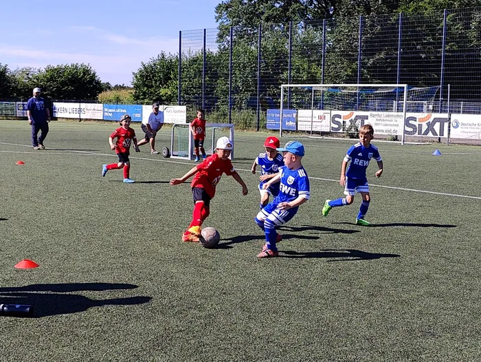 Junge Fußballspieler in roten und blauen Trikots während eines Spiels auf einem Kunstrasenplatz. Im Hintergrund eine Bande mit Werbeaufdrucken und Bäume.