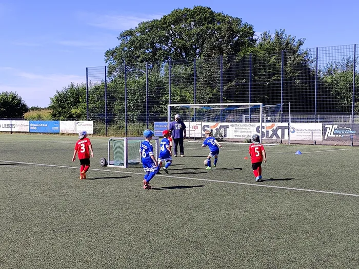 Fußballspieler in blauen und roten Trikots spielen auf einem grünen Kunstrasenplatz. Im Hintergrund eine Zaunbegrenzung und Werbebanner.