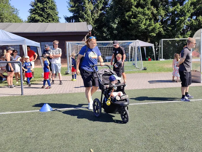 Eine Frau schiebt einen Kinderwagen über einen Kunstrasenplatz, während im Hintergrund Zuschauer und Fußballtore zu sehen sind. Im Hintergrund steht ein Zelt und weitere Personen beobachten das Geschehen.