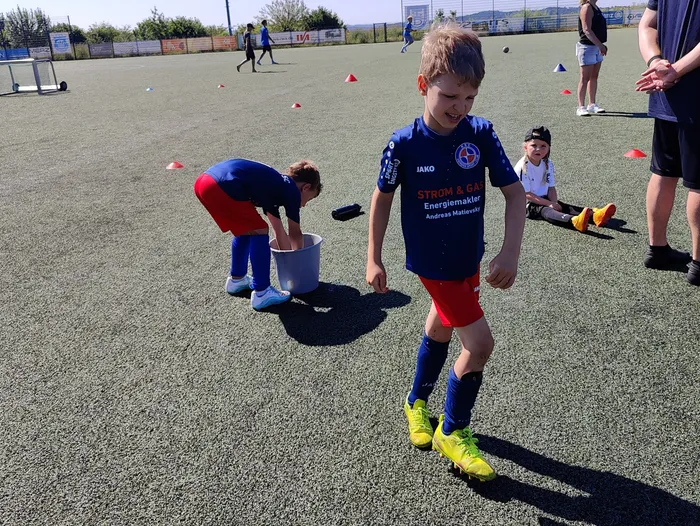 Ein Jugendlicher in Fußballkleidung läuft auf einem grünen Kunstrasenplatz, während ein zweiter Jugendlicher einen Eimer aufhebt. Im Hintergrund trainieren weitere Spieler.