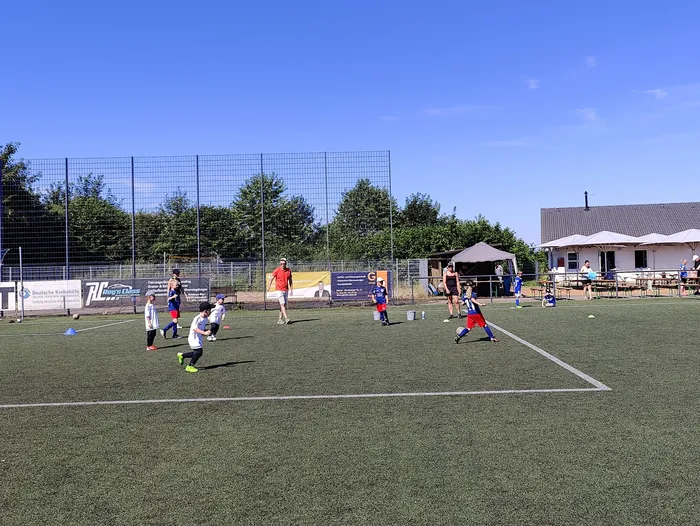 Fußballspieler in blauen und roten Trikots während eines Spiels auf einem Kunstrasenplatz. Im Hintergrund ein Zaun, Zuschauer und ein Gebäude.