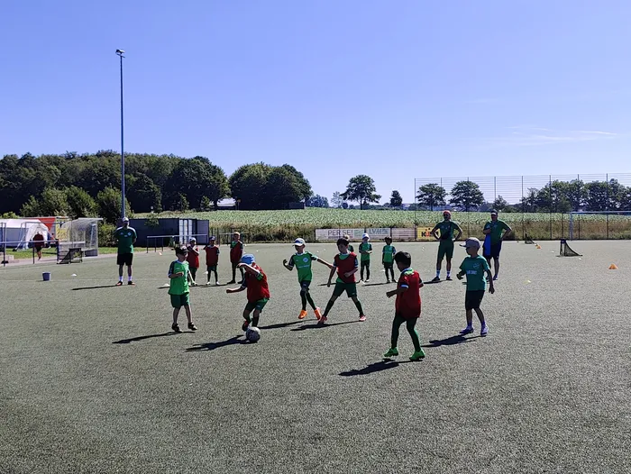 Eine Gruppe Jugendlicher in rot und grün spielt Fußball auf einem Kunstrasenplatz. Im Hintergrund sind Bäume und ein blau-gelber Werbebanner zu sehen.
