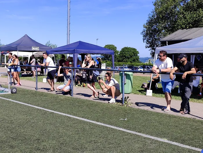 Zuschauer stehen und sitzen an einer Absperrung und beobachten eine Sportveranstaltung auf einem grünen Kunstrasenplatz. Im Hintergrund sind mehrere blaue Pavillons und Bäume zu sehen.