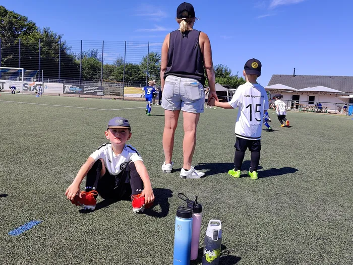 Eine Frau führt zwei Kinder in Fußballtrikots über einen Fußballplatz. Im Vordergrund kauert ein Kind mit Handschuhen und schaut in die Kamera. Im Hintergrund sind weitere spielende Kinder und Zuschauer zu sehen.
