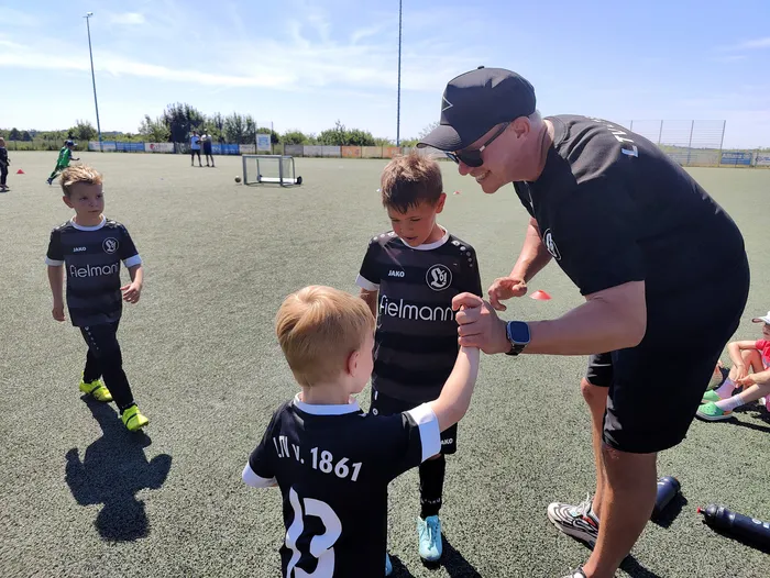 Ein Trainer spricht mit jungen Fußballspielern auf einem Sportplatz. Die Kinder tragen Trikots mit dem Vereinslogo und der Aufschrift 