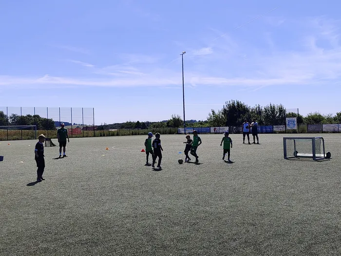 Eine Gruppe Jugendlicher spielt Fußball auf einem Kunstrasenplatz. Im Hintergrund sind ein Zaun, Bäume und ein Fußballtor zu sehen.