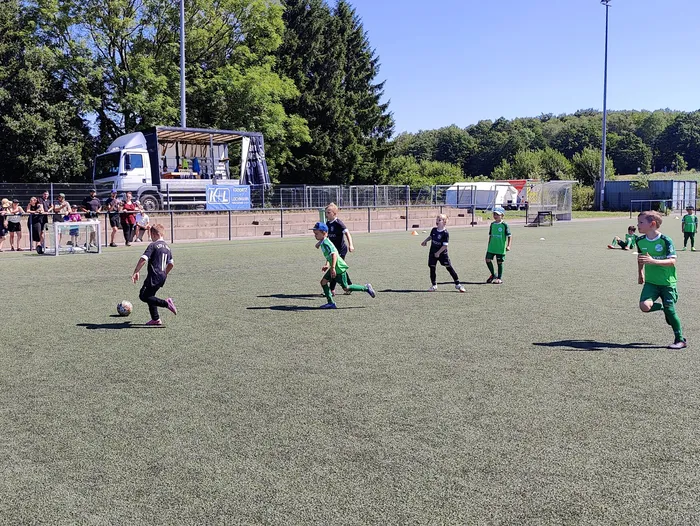 Fußballspieler in Schwarz und Grün auf einem grünen Rasenfeld während eines Turniers. Im Hintergrund stehen Zuschauer und ein Imbisswagen.