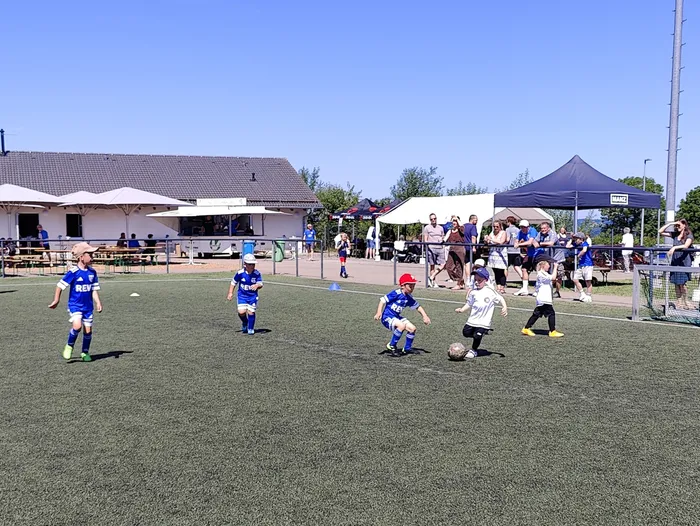 Junge Fußballspieler in blauen und weißen Trikots während eines Spiels auf einem Kunstrasenplatz. Im Hintergrund ein Vereinsheim und Zuschauer unter einem Pavillon.