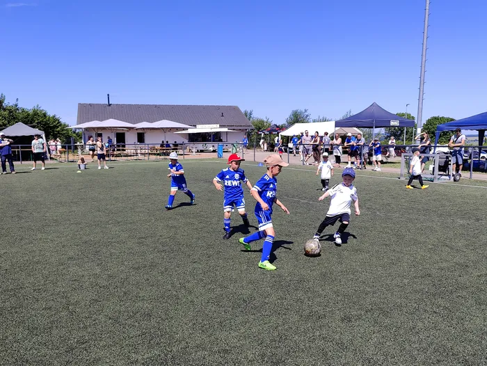 Junge Fußballspieler in blauen und weißen Trikots kämpfen um den Ball auf einem grünen Kunstrasenplatz. Im Hintergrund stehen Zuschauer und ein Gebäude mit einem Pavillon.