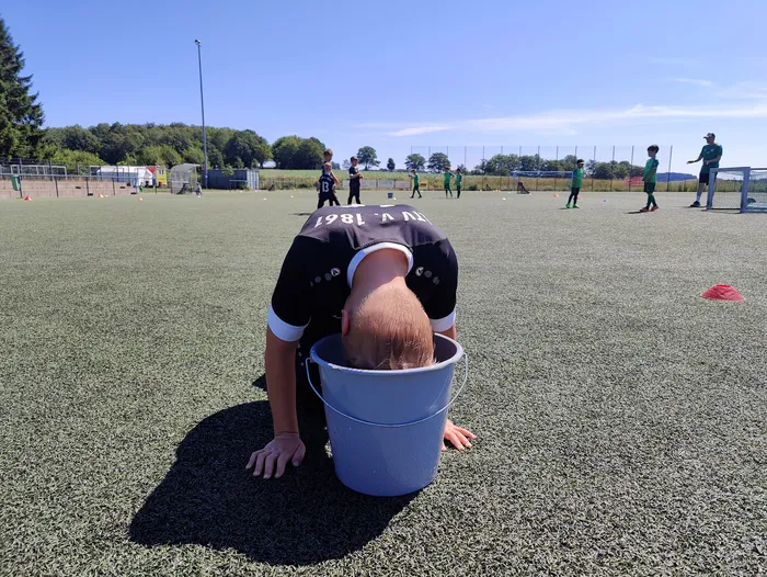 Ein Fußballspieler mit dem Kopf in einem Eimer steht auf einem grünen Kunstrasenplatz. Im Hintergrund sind weitere Spieler und Trainer zu sehen.