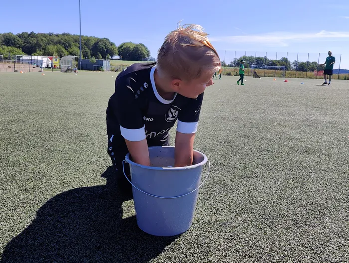 Ein Junge in Fußballkleidung kniet auf einem grünen Kunstrasenplatz und hält einen Eimer vor sich. Im Hintergrund sind weitere Spieler und Trainingshütchen zu sehen.
