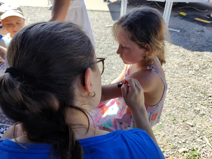 Eine Frau bemalt dem kleinen Mädchen einen Arm. Im Hintergrund ist ein Kind mit einer Baseballkappe zu sehen.